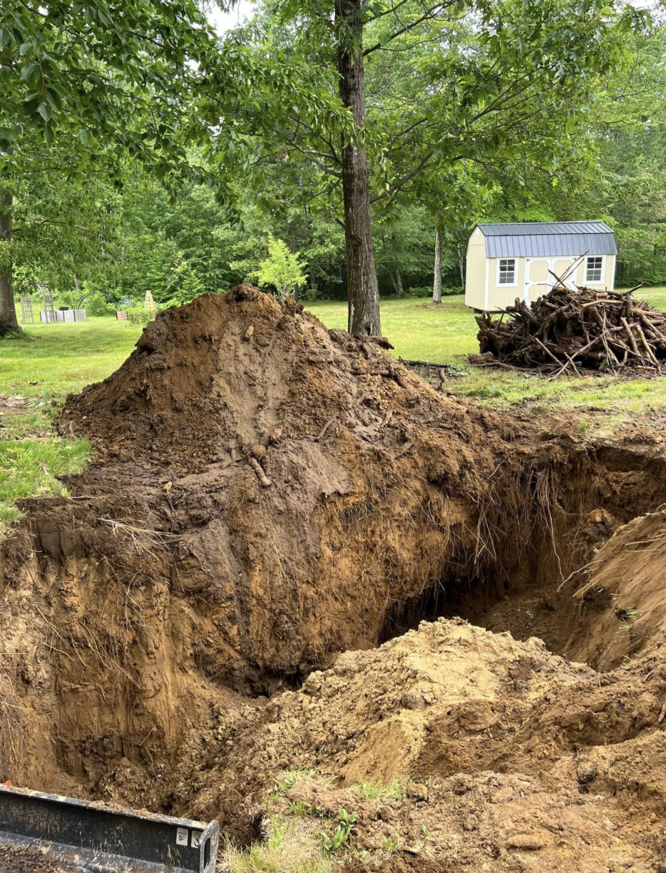 shutterstock_2123972297 (1) Excavation in a backyard with a view of trees, a shed, and a pile of old stumps in the background.